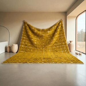 Large handmade Moroccan high-pile shag rug in saturated Thai curry mustard-gold, displayed vertically against a white brick wall. Surface features a tone-on-tone square grid pattern formed through pile variation — raised mustard squares separated by recessed ivory cream channel lines across the entire field. Dense, plush, curly wool pile texture throughout. Knotted braided ivory fringe along the top hem edge and long hand-tied ivory fringe along the bottom edge.