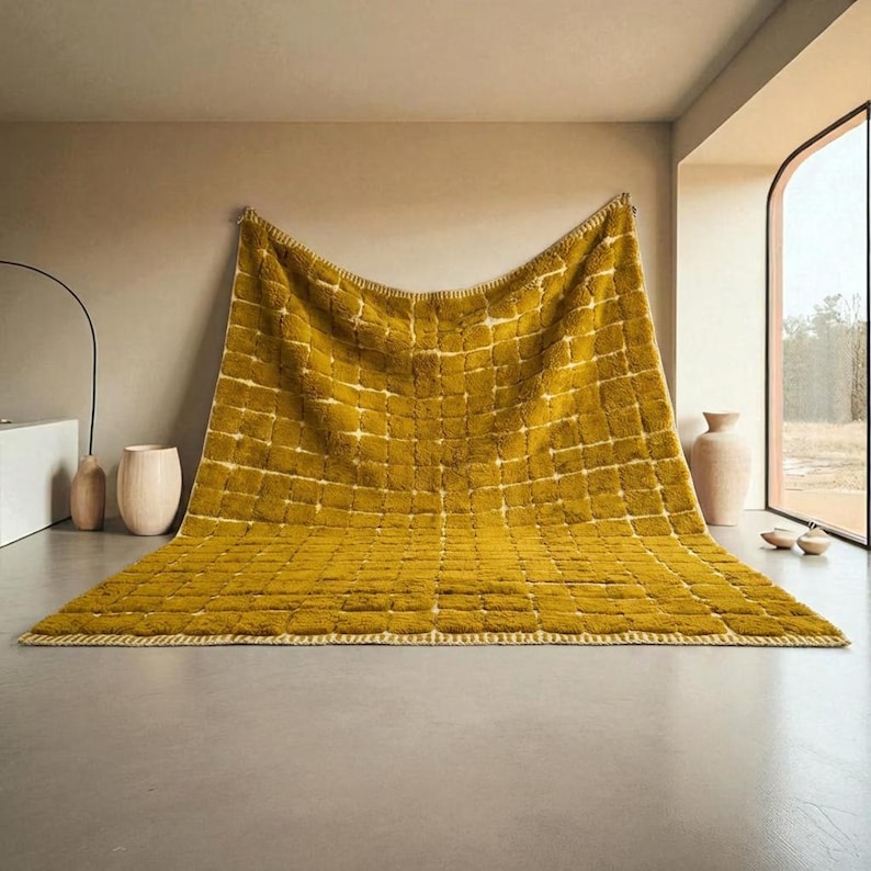 Large handmade Moroccan high-pile shag rug in saturated Thai curry mustard-gold, displayed vertically against a white brick wall. Surface features a tone-on-tone square grid pattern formed through pile variation — raised mustard squares separated by recessed ivory cream channel lines across the entire field. Dense, plush, curly wool pile texture throughout. Knotted braided ivory fringe along the top hem edge and long hand-tied ivory fringe along the bottom edge.