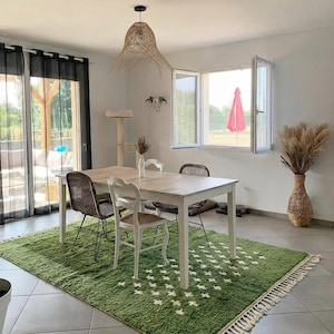 A plush handwoven Moroccan Berber rug in a rich moss green. The thick wool carpet features a solid dark green band near the top and a lighter mossy green base beautifully adorned with small, cream-colored geometric diamond motifs at the bottom. It is draped elegantly against a white painted brick wall next to a gold accent table with trailing ivy, perfectly capturing a warm, earthy, and organic modern interior design aesthetic.