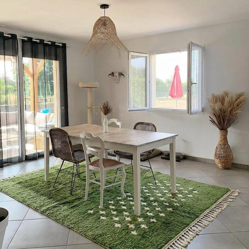 A plush handwoven Moroccan Berber rug in a rich moss green. The thick wool carpet features a solid dark green band near the top and a lighter mossy green base beautifully adorned with small, cream-colored geometric diamond motifs at the bottom. It is draped elegantly against a white painted brick wall next to a gold accent table with trailing ivy, perfectly capturing a warm, earthy, and organic modern interior design aesthetic.