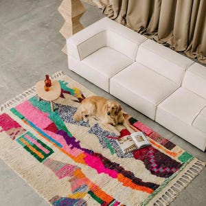 A vibrant, high-pile handmade Moroccan wool rug displayed in a bright industrial studio against a white brick wall. The rug features an eclectic, abstract tribal design with bold splashes of neon pink, teal green, orange, royal blue, and charcoal grey on a natural cream base. The patterns include geometric shapes, abstract figures, and spontaneous color blocking. The rug showcases a thick, plush shaggy texture and authentic hand-knotted cream tassels at the bottom.