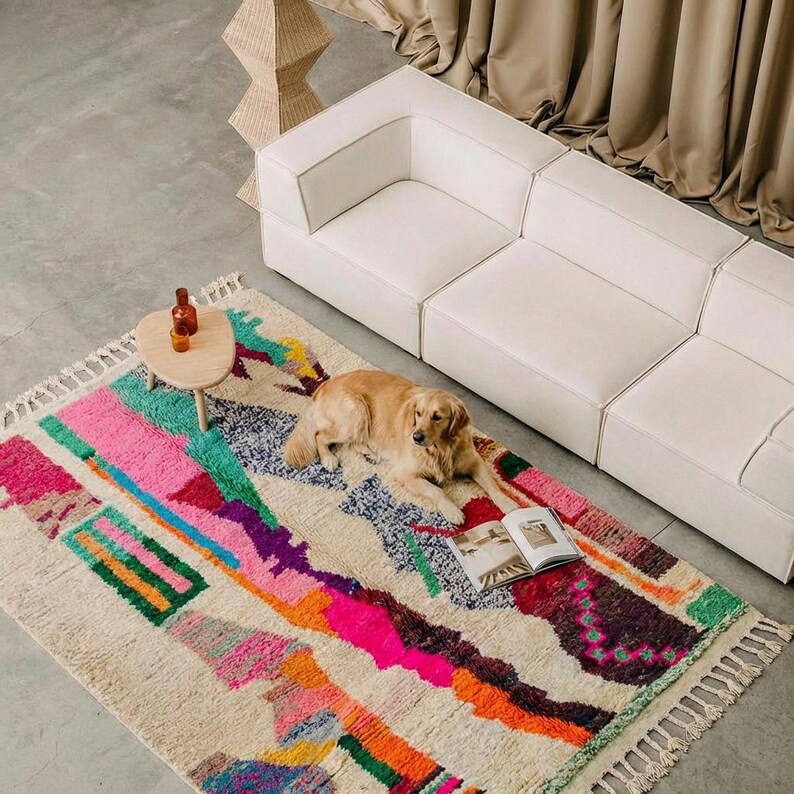 A vibrant, high-pile handmade Moroccan wool rug displayed in a bright industrial studio against a white brick wall. The rug features an eclectic, abstract tribal design with bold splashes of neon pink, teal green, orange, royal blue, and charcoal grey on a natural cream base. The patterns include geometric shapes, abstract figures, and spontaneous color blocking. The rug showcases a thick, plush shaggy texture and authentic hand-knotted cream tassels at the bottom.