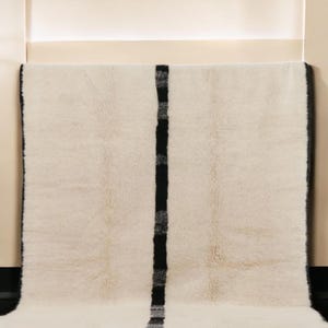 Handmade Moroccan wool rug featuring a minimalist black and white design. Ethically crafted by Berber artisans in Morocco using 100% natural wool. Thick, plush, and ultra-soft, this Beni Ourain-style carpet is perfect for modern, Scandinavian, and bohemian interiors. Ideal for living rooms, bedrooms, and luxury home decor. Available in custom sizes. Authentic tribal craftsmanship with eco-friendly materials. Free worldwide shipping with FedEx Express.