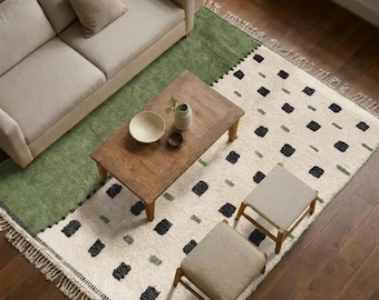 May include: A rectangular area rug featuring a cream-coloured field with a pattern of black and green rectangular tufts, finished with fringe on the ends. The rug is positioned under a beige sofa, a wooden coffee table, and two beige stools.