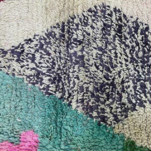 A vibrant, high-pile handmade Moroccan wool rug displayed in a bright industrial studio against a white brick wall. The rug features an eclectic, abstract tribal design with bold splashes of neon pink, teal green, orange, royal blue, and charcoal grey on a natural cream base. The patterns include geometric shapes, abstract figures, and spontaneous color blocking. The rug showcases a thick, plush shaggy texture and authentic hand-knotted cream tassels at the bottom.