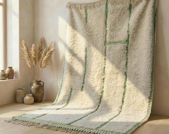 May include: A large, rectangular, off-white shag rug featuring a series of vertical and horizontal thin green lines. The rug has a thick, textured pile and is finished with a fringe of knotted yarn along the bottom edge, creating a soft appearance.