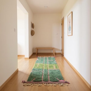 Puede incluir: Una alfombra rectangular con un patrón geométrico en tonos verdes y rosas en un pasillo. La alfombra está colocada sobre un suelo de madera y tiene flecos en los extremos. Es un elemento decorativo para el hogar.