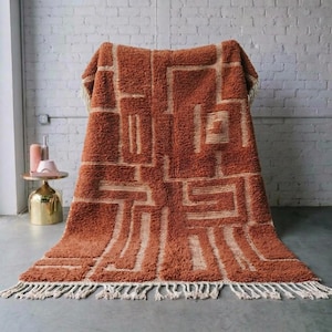May include: A rust-colored, geometric patterned rug with a cream-colored design. The rug has a shaggy texture and a fringe along the bottom edge. The rug is draped over a chair against a white brick wall.