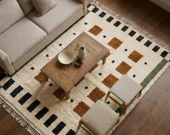 May include: A cream-coloured area rug featuring a geometric pattern of brown and black squares and rectangles, finished with fringe on two ends. The rug is positioned under a wooden coffee table, two small stools, and a beige sofa with a throw pillow.