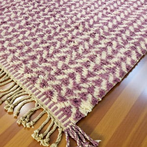 Close-up of a Wistful Mauve Moroccan wool rug handwoven from natural wool by Berber artisans. The rug features a soft mauve and cream color palette with a beautiful geometric pattern and traditional fringes. This thick and plush Moroccan carpet adds warmth, softness, and artisanal charm to living rooms, bedrooms, or boho-chic interiors.