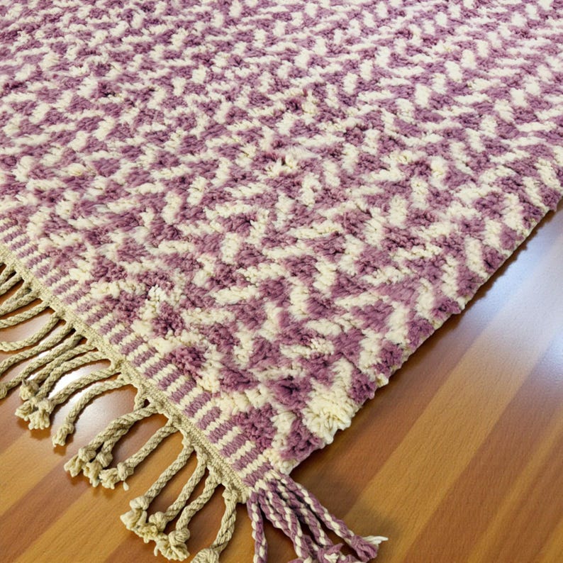 Close-up of a Wistful Mauve Moroccan wool rug handwoven from natural wool by Berber artisans. The rug features a soft mauve and cream color palette with a beautiful geometric pattern and traditional fringes. This thick and plush Moroccan carpet adds warmth, softness, and artisanal charm to living rooms, bedrooms, or boho-chic interiors.