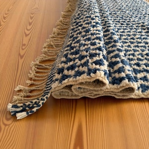 May include: A folded area rug featuring a repeating blue and cream geometric pattern with a textured pile. The rug is finished with long, cream-coloured twisted fringe tassels along the edge, creating a decorative border on the textile piece.