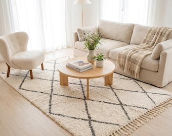 May include: A cream-coloured area rug featuring a dark grey diamond grid pattern and fringed edges. The rug is placed in a room with a beige sofa, a matching armchair, a round wooden coffee table, and a floor lamp with a white shade.