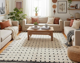 May include: A living room with a cream sofa, patterned rug, and wooden coffee table. The rug is white with black square patterns and a dark border. Cushions in various colours and textures decorate the sofa. The room has a bohemian style.