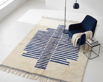 Può includere: Un tappeto rettangolare con base color crema e un motivo geometrico centrale a strisce orizzontali blu navy. Il tappeto presenta frange alle estremità ed è posizionato sotto una poltrona blu scuro e una lampada da terra.