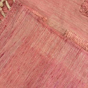 Handwoven Moroccan wool area rug in soft solid pink with a plush, high-pile texture and natural cream wool fringes. Made by Amazigh artisans in the Atlas Mountains, this minimalist Beni Ourain-style rug brings warmth, softness, and authentic Moroccan craftsmanship to modern boho or minimalist interiors.