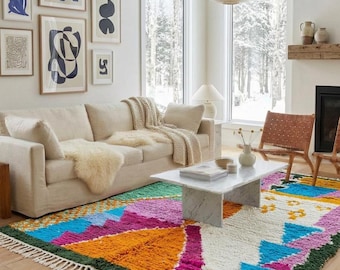 Può includere: Un tappeto rettangolare con un vivace motivo geometrico nei colori rosa acceso, arancione, blu, verde e bianco, rifinito con frange bianche alle estremità. Il tappeto è posizionato in un soggiorno con divano beige, tavolino in marmo e quadri.