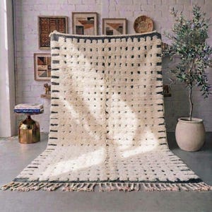Handmade Moroccan wool rug featuring a warm ivory background with gray borders, short linear accents, and small irregular dotted motifs arranged across the surface. Individually hand-knotted by skilled Amazigh artisans in the Atlas Mountains using 100% natural sheep wool, its thick plush high pile adds softness, warmth, and authentic handcrafted texture. Finished with natural ivory fringes, this refined rug organic modern, Japandi, minimalist, Scandinavian, contemporary, and neutral interiors.