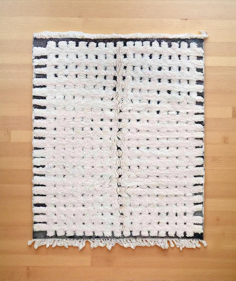 Handmade Moroccan wool rug featuring a warm ivory background with gray borders, short linear accents, and small irregular dotted motifs arranged across the surface. Individually hand-knotted by skilled Amazigh artisans in the Atlas Mountains using 100% natural sheep wool, its thick plush high pile adds softness, warmth, and authentic handcrafted texture. Finished with natural ivory fringes, this refined rug organic modern, Japandi, minimalist, Scandinavian, contemporary, and neutral interiors.