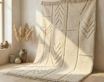 May include: A cream-coloured area rug with a high-pile texture and a geometric pattern of raised chevron lines. The rug features a fringed edge along the top and bottom, showcasing a soft, shaggy appearance with vertical panels of repeating arrow motifs.