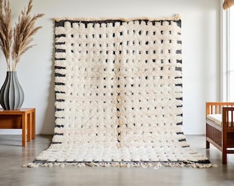 May include: A large, rectangular rug with a cream-colored background and a pattern of black dots and lines. The rug has a thick, textured pile and a fringe along the bottom edge. The rug is hanging on a white wall.