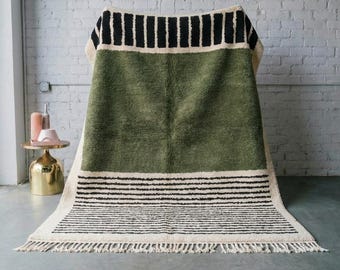 May include: A green and cream-colored rug with black stripes and fringe. The rug has a large green central section, bordered by cream-colored sections with black stripes. The bottom edge features a cream-colored fringe.