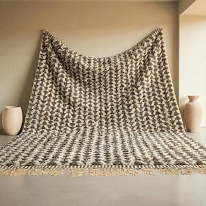 Könnte beinhalten: Eine große, gewebte Decke mit einem sich wiederholenden geometrischen Muster in Braun- und Cremetönen. Die Decke hat einen Fransenrand und ist über eine Wand und einen Boden drapiert. Das Gesamtthema ist Wohnkultur.