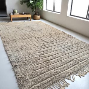 Puede incluir: Alfombra rectangular beige con un patrón geométrico de rombos en el centro. La alfombra tiene una superficie texturizada con líneas horizontales y flecos en los bordes. La alfombra está colocada sobre un suelo blanco.