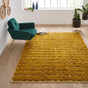 Large handmade Moroccan high-pile shag rug in saturated Thai curry mustard-gold, displayed vertically against a white brick wall. Surface features a tone-on-tone square grid pattern formed through pile variation — raised mustard squares separated by recessed ivory cream channel lines across the entire field. Dense, plush, curly wool pile texture throughout. Knotted braided ivory fringe along the top hem edge and long hand-tied ivory fringe along the bottom edge.