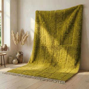 Puede incluir: Una alfombra grande de color verde oliva con un patrón geométrico texturizado. La alfombra se muestra contra una pared blanca y tiene flecos en el borde inferior. La alfombra está hecha de un material suave y se coloca sobre un suelo de madera clara. Dimensiones: aprox. 200 cm x 140 cm.