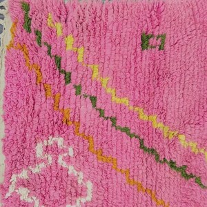 A plush, handwoven Moroccan Boujad rug featuring a vibrant hot pink base adorned with neon orange crosses, white diamonds, and green zigzag geometric patterns. The brightly colored wool carpet is draped elegantly against a white painted brick wall next to a gold accent table with pink pillar candles, perfectly capturing a playful maximalist and dopamine decor aesthetic.