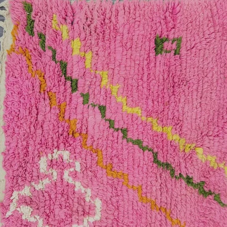 A plush, handwoven Moroccan Boujad rug featuring a vibrant hot pink base adorned with neon orange crosses, white diamonds, and green zigzag geometric patterns. The brightly colored wool carpet is draped elegantly against a white painted brick wall next to a gold accent table with pink pillar candles, perfectly capturing a playful maximalist and dopamine decor aesthetic.