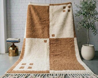 May include: A rectangular area rug featuring a large-scale checkerboard pattern in alternating blocks of cream and tan. The rug has a thick, shaggy pile texture and is finished with a row of knotted fringe along the bottom edge.