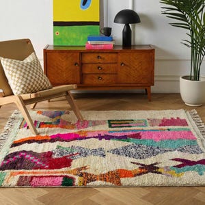 Colorful Moroccan wool rug in a boho living room, featuring an ivory base with abstract patchwork blocks in pink, orange, green, burgundy, black, yellow, and mint, plus playful linear motifs and braided ivory fringes. The rug has a plush high pile and hand-knotted texture, styled under a round rattan coffee table with a cream sofa, earthy pillows, woven pouf, plants, macramé wall art, and warm natural light. A vibrant Azilal rug for boho, eclectic, kids room, and creative interiors.