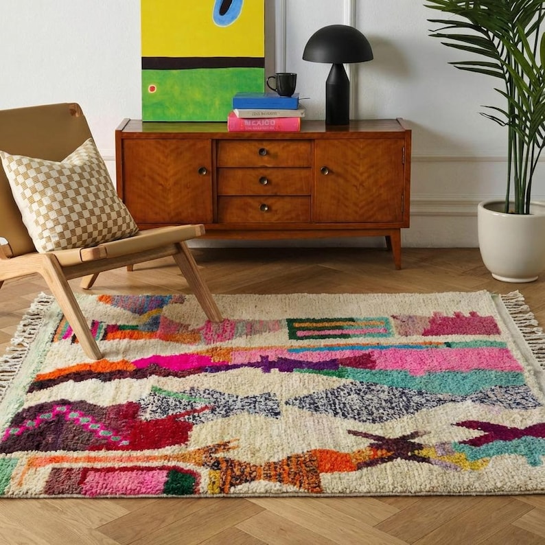 Colorful Moroccan wool rug in a boho living room, featuring an ivory base with abstract patchwork blocks in pink, orange, green, burgundy, black, yellow, and mint, plus playful linear motifs and braided ivory fringes. The rug has a plush high pile and hand-knotted texture, styled under a round rattan coffee table with a cream sofa, earthy pillows, woven pouf, plants, macramé wall art, and warm natural light. A vibrant Azilal rug for boho, eclectic, kids room, and creative interiors.