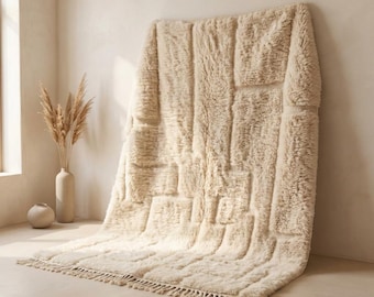 May include: A large, cream-colored rug with a geometric pattern and a fringed edge. The rug is made of a thick, textured material and is leaning against a wall. The rug is approximately 60 inches wide and 80 inches long.