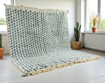 May include: A large, rectangular rug with a repeating geometric pattern in shades of blue and cream. The rug has a textured surface and is trimmed with cream-coloured fringe along the edges. The rug is displayed against a white wall.