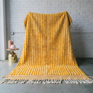 May include: A yellow and white striped rug with a textured surface and fringe detail. The rug is draped over a chair, showcasing its vertical stripe pattern. The rug's colour palette includes shades of yellow and white.