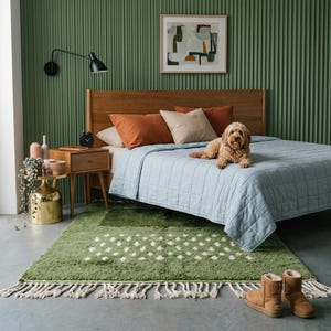 A plush handwoven Moroccan Berber rug in a rich moss green. The thick wool carpet features a solid dark green band near the top and a lighter mossy green base beautifully adorned with small, cream-colored geometric diamond motifs at the bottom. It is draped elegantly against a white painted brick wall next to a gold accent table with trailing ivy, perfectly capturing a warm, earthy, and organic modern interior design aesthetic.