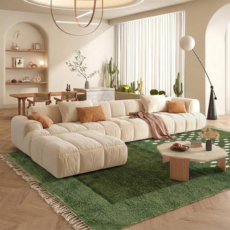 A plush handwoven Moroccan Berber rug in a rich moss green. The thick wool carpet features a solid dark green band near the top and a lighter mossy green base beautifully adorned with small, cream-colored geometric diamond motifs at the bottom. It is draped elegantly against a white painted brick wall next to a gold accent table with trailing ivy, perfectly capturing a warm, earthy, and organic modern interior design aesthetic.
