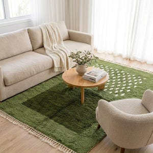Handmade Moroccan Boujaad rug in soft sage green with scattered ivory white dot motifs across the lower field. Thick plush shaggy wool pile with natural white knotted fringe. Styled in a serene minimalist room with a woman holding a newborn baby in a linen chair, a golden retriever lying on the rug, white curtains and built-in ceramic shelf display. Hand-knotted by Berber artisans in Marrakech, Morocco. A gentle, calming statement rug for nurseries, family rooms and boho interiors.