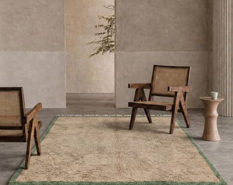 Può includere: Un tappeto rettangolare con un centro beige testurizzato, un sottile bordo verde e frange color bianco sporco sulle estremità corte. Due poltrone in legno con schienali e sedute in paglia di Vienna sono posizionate ai lati del tappeto.
