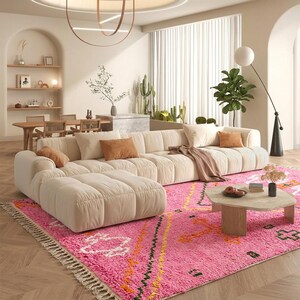A plush, handwoven Moroccan Boujad rug featuring a vibrant hot pink base adorned with neon orange crosses, white diamonds, and green zigzag geometric patterns. The brightly colored wool carpet is draped elegantly against a white painted brick wall next to a gold accent table with pink pillar candles, perfectly capturing a playful maximalist and dopamine decor aesthetic.