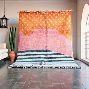 Può includere: Un grande tappeto colorato con un design astratto. La sezione superiore è arancione con pois bianchi, che sfuma in rosa, poi in strisce blu e bianche nella parte inferiore. Il tappeto ha un aspetto strutturato e tessuto, con un bordo sfrangiato.
