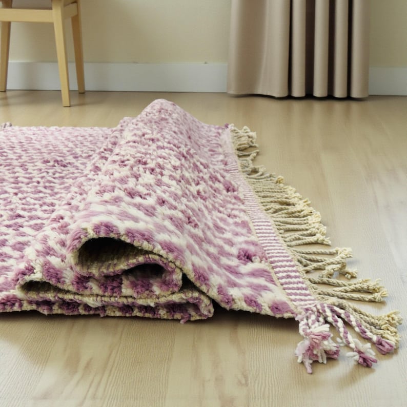 Close-up of a Wistful Mauve Moroccan wool rug handwoven from natural wool by Berber artisans. The rug features a soft mauve and cream color palette with a beautiful geometric pattern and traditional fringes. This thick and plush Moroccan carpet adds warmth, softness, and artisanal charm to living rooms, bedrooms, or boho-chic interiors.
