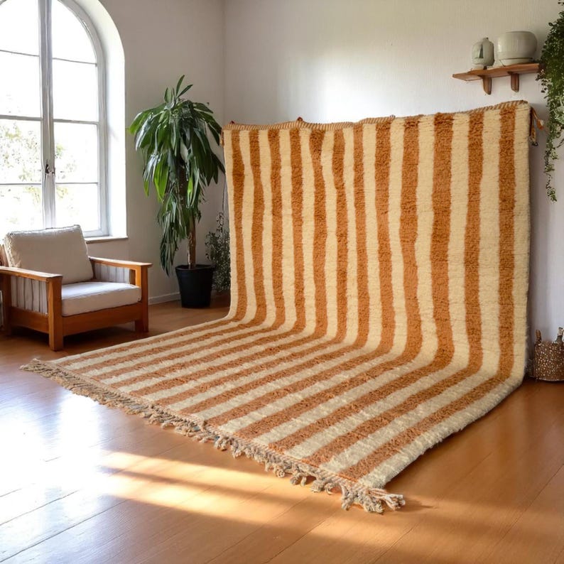 Elegant handwoven Moroccan rug with vertical stripes in Cameo Rose and soft white. Perfect for boho, coastal, minimalist, or Japandi-inspired spaces. Made from natural wool, this artisan piece blends texture and comfort for your living room, bedroom, or entryway. A modern pastel rug with warm earthy tones and timeless Amazigh character. Custom sizes available.