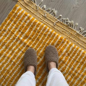 May include: A close-up of a yellow and white striped rug with a textured surface and a fringed border. Brown slippers rest on the rug. White trousers are visible.