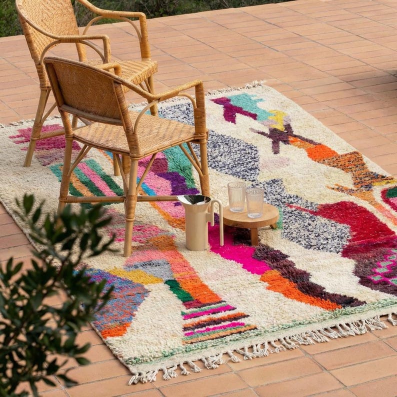 A vibrant, high-pile handmade Moroccan wool rug displayed in a bright industrial studio against a white brick wall. The rug features an eclectic, abstract tribal design with bold splashes of neon pink, teal green, orange, royal blue, and charcoal grey on a natural cream base. The patterns include geometric shapes, abstract figures, and spontaneous color blocking. The rug showcases a thick, plush shaggy texture and authentic hand-knotted cream tassels at the bottom.