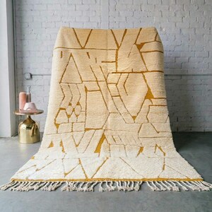Handmade Moroccan Beni Ourain wool rug in natural ivory with bold mustard gold freehand diamond and geometric line composition. Irregular angular mustard lines and diamond forms are scattered asymmetrically across a thick shaggy ivory pile creating a spontaneous abstract pattern. Knotted cream fringe along the bottom edge. Styled flat on oak wood flooring in a bright boho living room with plants, rattan, and macrame.