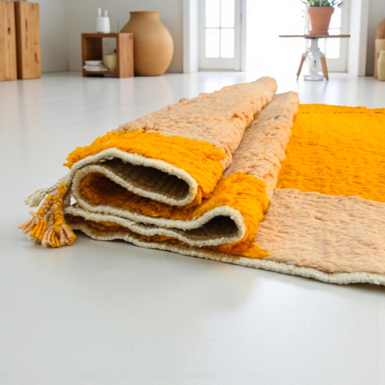 Handmade Moroccan rug in bold golden yellow and soft beige with a striking abstract black line design. This Berber wool carpet is woven by skilled artisans from Morocco, combining traditional craftsmanship with modern style. Its vibrant color contrast and unique pattern make it a perfect statement piece for living rooms, bedrooms, or boho-inspired spaces. Luxurious, eco-friendly, and durable, this Moroccan rug adds artisanal warmth and contemporary elegance to any home.